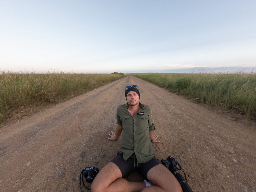 Portrait of photographer Michal Dzikowski sitting on a remote dirt road in the Tanzanian savannah surrounded by tall grass while working on hospitality and safari lodge photography projects.