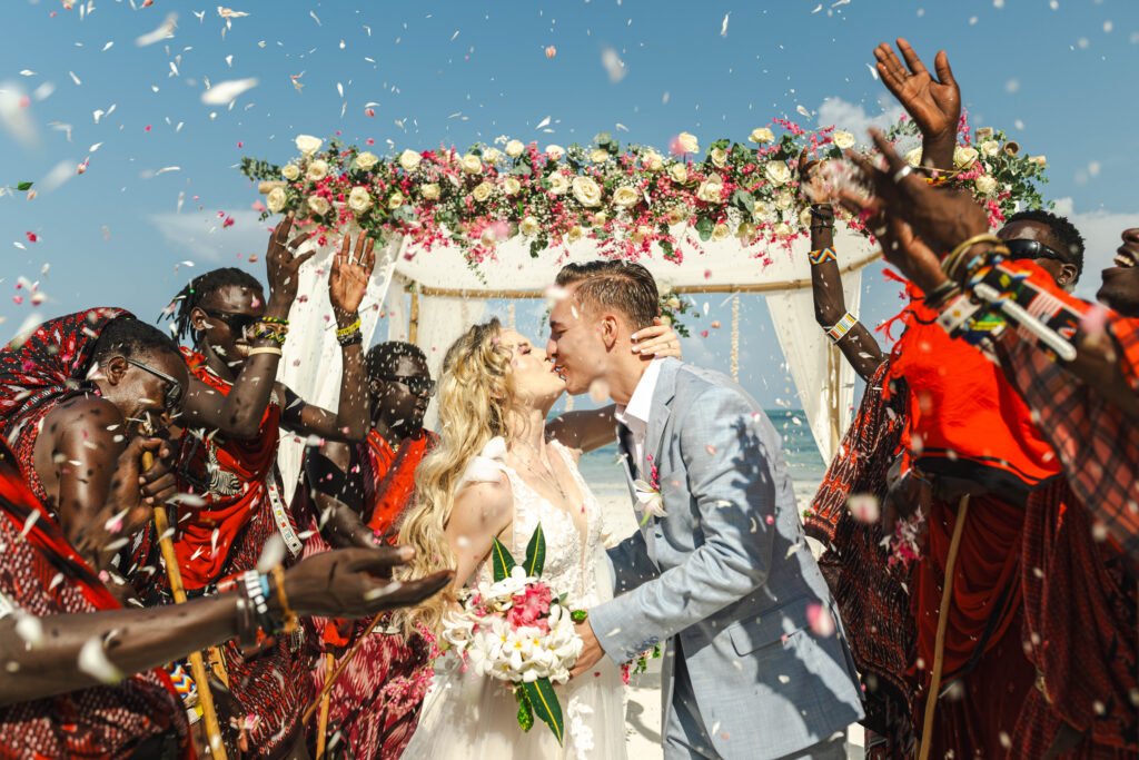 Bride and groom kissing during a Zanzibar beach wedding ceremony surrounded by Maasai dancers and flower petals.